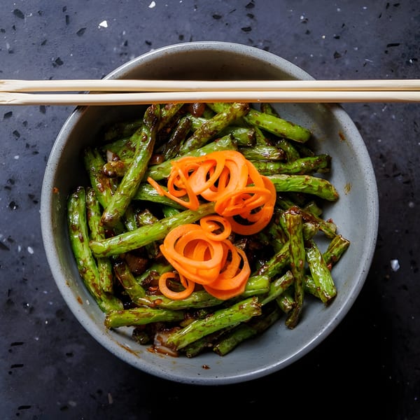 A bowl of green beans with sauce and carrot spirals, with chopsticks resting on top.