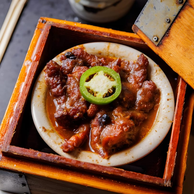 Asian braised pork belly in a white bowl, garnished with a green jalapeño slice, served in a wooden box.